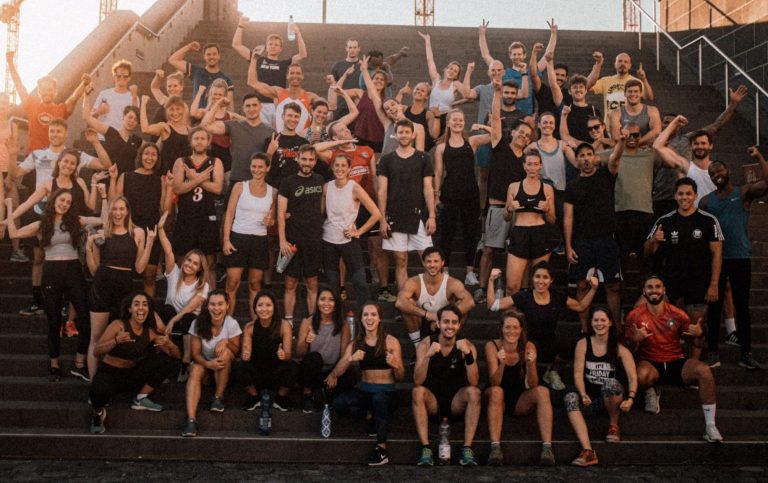 Aufnahme der Social Workout Crew bei Dämmerung im Ostendpark: Eine energiegeladene Gruppe von etwa 50 Sportlern in bunter Trainingskleidung versammelt sich auf der Treppe. Im Hintergrund spiegeln sich warme Sonnenuntergangsfarben im Glasturm der EZB, mit sichtbarer Deutschherrnbrücke und rotem Baukran. Vorne leuchten Laternen und eine Fahne mit abstraktem Muster.