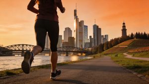 Laufbegeisterte Frankfurt‑Läuferin vor Frankfurter Skyline mit Mainufer, Stadtwald, Niddapark‑Wegen und Lohrberg‑Weinbergen – Übersichts‑Beitragsbild für Artikel ‚Laufen in Frankfurt: Die 5 besten Laufstrecken‘.