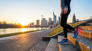 Nahaufnahme des Unterkörpers eines Läufers, der an einem Flussufer in Frankfurt bei Sonnenaufgang eine Treppe hinuntersteigt. Am rechten Fuß, der auf einer Stufe aufsetzt, ist ein rot leuchtender, pulsierender Kreis um die Ferse zu sehen, der Schmerz oder eine Verletzung wie Plantarfasziitis symbolisiert. Im Hintergrund die Skyline von Frankfurt am Main